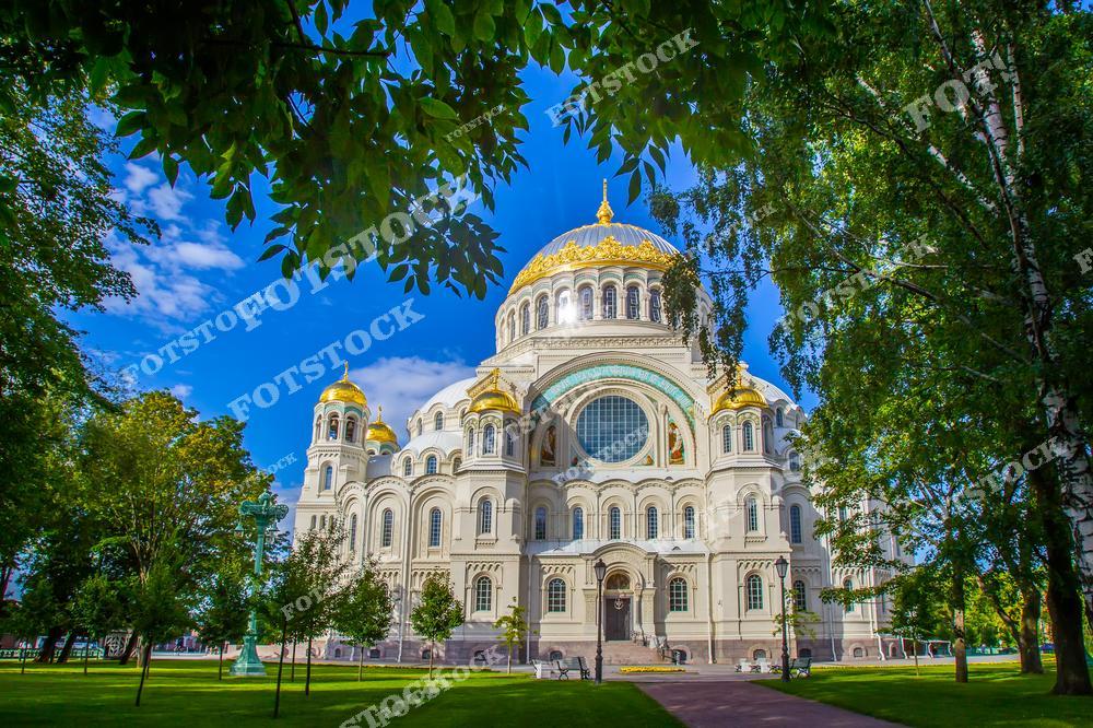 На фотографии изображен Морской Никольский собор, окружённый зеленью деревьев в летнем парке Кронштадта. Его золочёные купола сверкают на солнце, а фасад украшен мозаиками и арками, отражающими русско-византийский стиль. Это значимое здание было возведено в память о моряках и является одной из главных достопримечательностей Кронштадта. Летний пейзаж с яркой растительностью подчёркивает гармоничное сочетание природы и архитектуры.